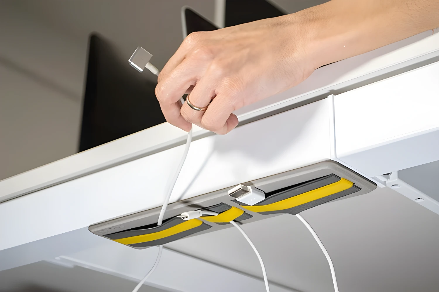 Organizador de cables adhesivo gris y amarillo en la parte inferior de un escritorio, sujetando cables y un adaptador USB.
