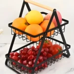 Estante frutero negro de dos niveles con asa de madera, lleno de naranjas, limón, uvas rojas y tomates en un mostrador blanco.