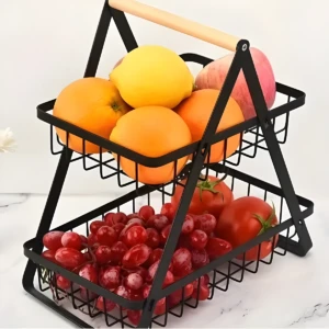 Estante frutero negro de dos niveles con asa de madera, lleno de naranjas, limón, uvas rojas y tomates en un mostrador blanco.