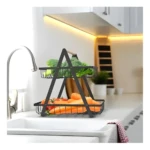 Cesta metálica negra de dos niveles con asa de madera, exhibiendo brócoli arriba y zanahorias y tomates abajo, sobre una encimera de cocina junto al lavaplatos.