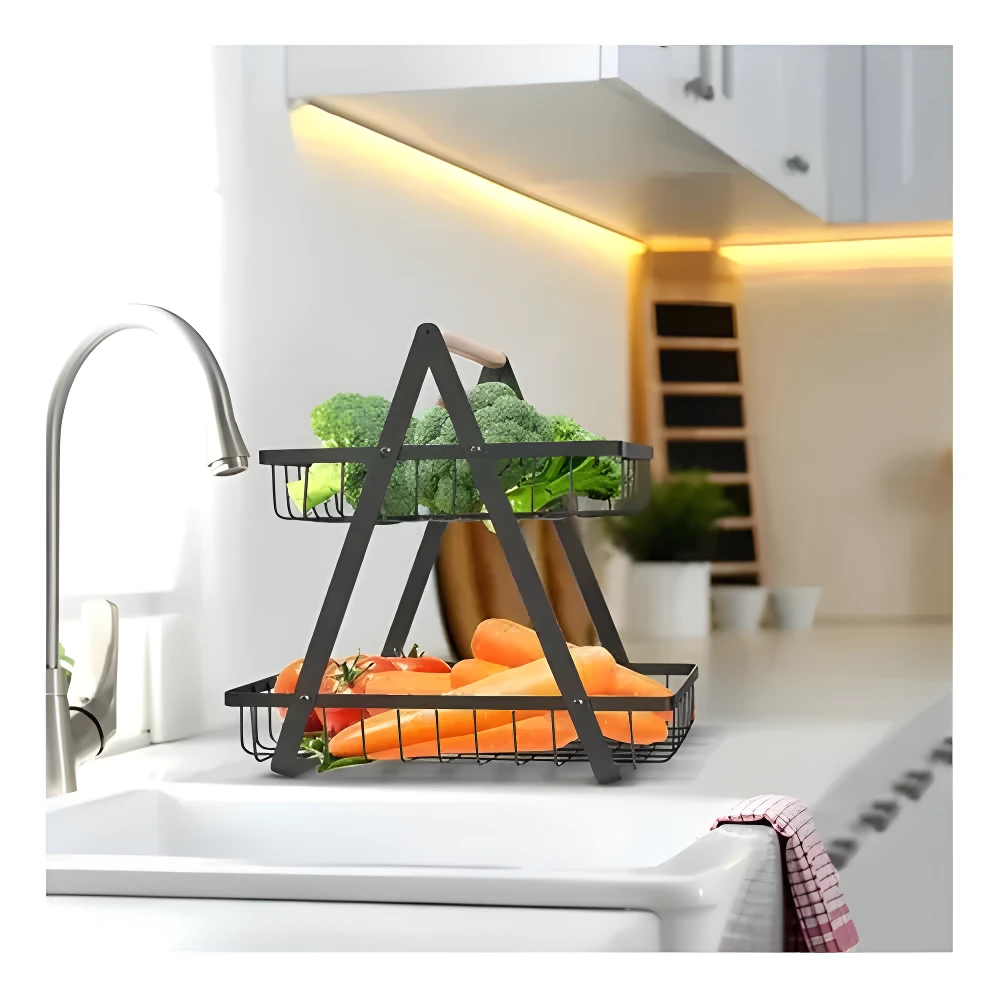 Cesta metálica negra de dos niveles con asa de madera, exhibiendo brócoli arriba y zanahorias y tomates abajo, sobre una encimera de cocina junto al lavaplatos.