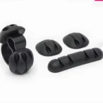 Juego de 4 organizadores de cables negros: uno tipo clip doble y tres soportes adhesivos planos para escritorio.