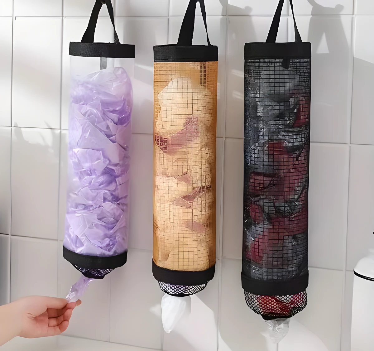 Tres dispensadores verticales de malla para bolsas plásticas: uno transparente con bolsas moradas, uno dorado y uno negro, colgados en pared de azulejos blancos.