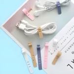 Organizadores de cable de silicona reutilizables en colores pastel: blanco, beige, gris y rosa, sujetando cables de audífonos y USB sobre una superficie con revista.