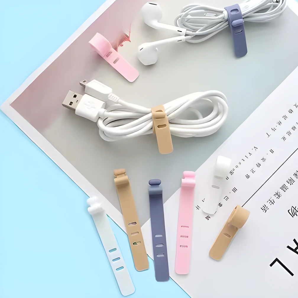 Organizadores de cable de silicona reutilizables en colores pastel: blanco, beige, gris y rosa, sujetando cables de audífonos y USB sobre una superficie con revista.