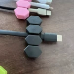 Organizador de cables magnético hexagonal, sujetando un cable USB-C negro, con otros cables rosa y gris al fondo sobre superficie marrón.