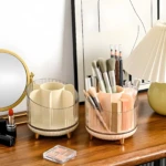 Dos organizadores redondos de acrílico y plástico, uno beige y otro rosado, exhibiendo brochas de maquillaje, lápices y labiales sobre una cómoda de madera.