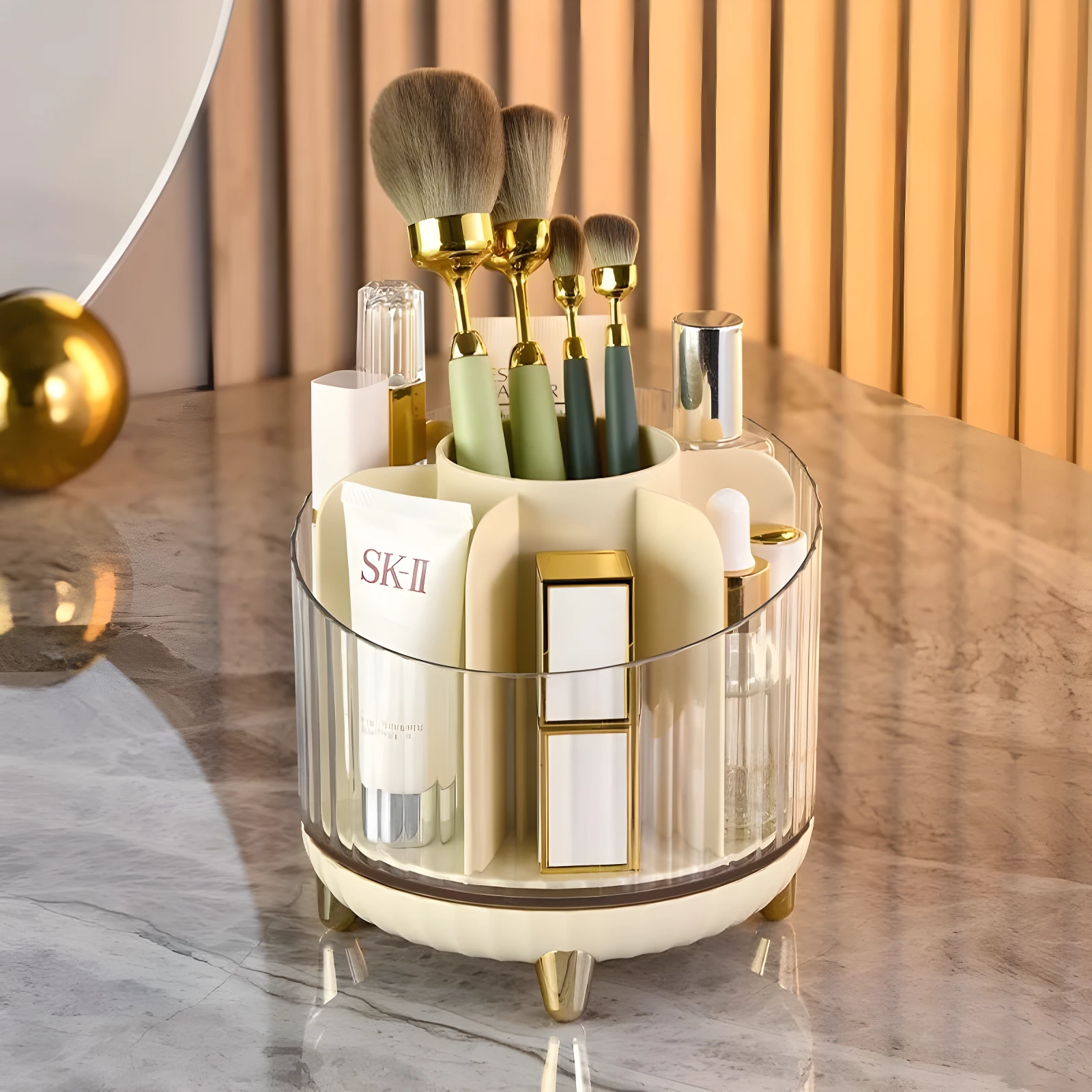Organizador giratorio de cosméticos transparente y crema, con patas doradas, exhibiendo brochas, crema SK-II y labiales.