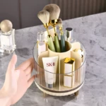 Organizador giratorio de maquillaje acrílico con divisiones, mostrando brochas, labial dorado y crema SK-II, con una mano al lado.
