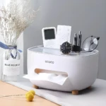 Organizador de escritorio blanco con dispensador de pañuelos y compartimentos para controles, bolígrafos y reloj.