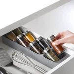 Organizador gris inclinado para cajón con frascos de especias de vidrio y tapas metálicas; mano sacando pimentón.