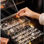 Joyero apilable acrílico transparente con compartimentos organizando collares, pulseras y anillos, mano colocando detalle floral.