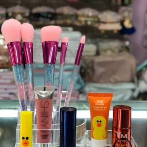 Organizador acrílico transparente con brochas de cerdas rosas, labial pato amarillo, brillo Victoria's Secret, crema de manos y esmalte rojo espejo.