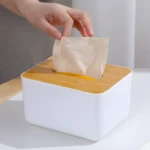 Dispensador de pañuelos blanco con tapa de bambú, mano sacando un pañuelo.