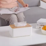 Dispensador de pañuelos blanco con tapa de bambú en mesa, persona leyendo al fondo