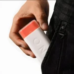 Organizador de pastillas de viaje blanco con tapa naranja, en mano cerca de pantalón negro con etiqueta Vogue.