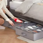 Organizador de zapatos gris bajo cama con compartimentos y tapa transparente, una persona guarda un zapato rojo.