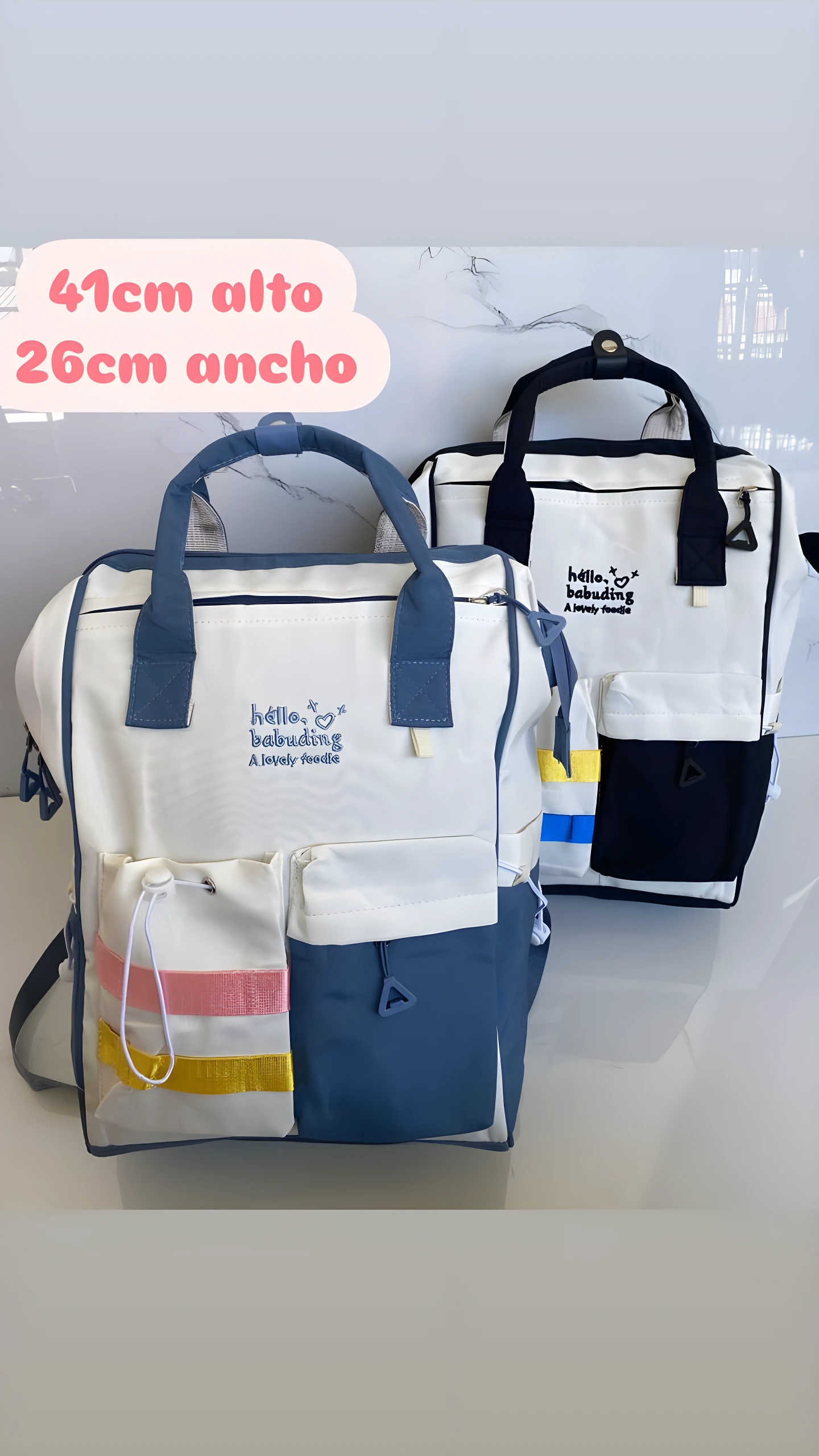 Pañalera morral blanca y azul, con dos compartimentos frontales y detalles a rayas, ideal para organizar artículos de bebé.