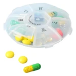 Pastillero organizador redondo transparente con 7 compartimentos para medicamentos, incluye pastillas amarillas y cápsula verde.