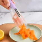 Pelador de vegetales lavanda con depósito transparente vaciando tiras de zanahoria sobre plato verde, junto a un limón.
