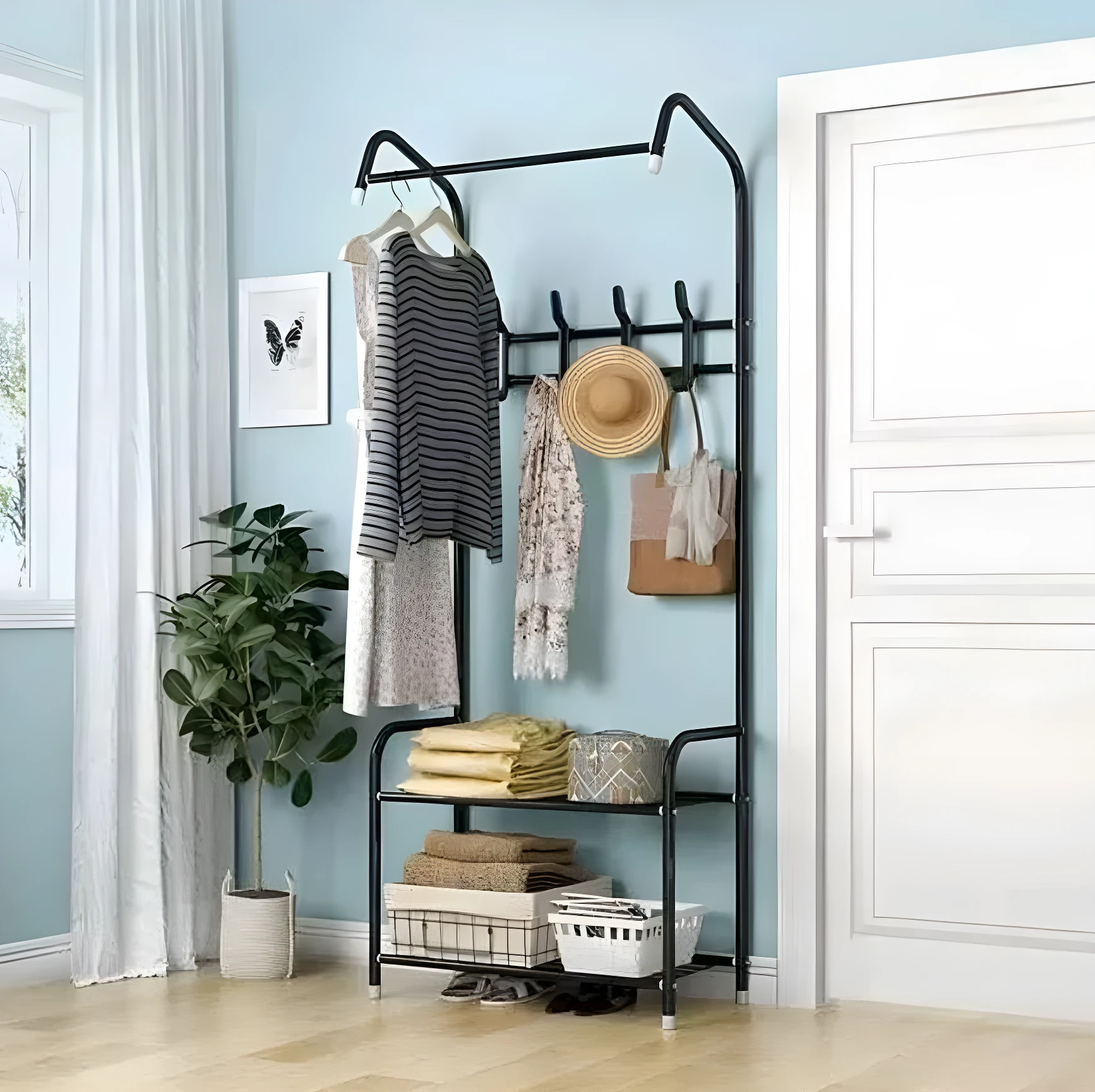 Perchero de pie negro metálico con barra superior y dos estantes inferiores, exhibiendo ropa, sombrero y bolsos en una entrada azul claro.