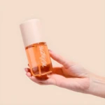 Perfume capilar y corporal Milagros, envase naranja transparente con atomizador, sostenido en mano con uñas pintadas.