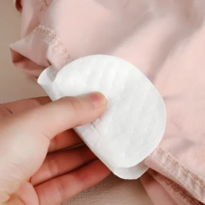 Mano sosteniendo parche blanco autoadhesivo para axilas sobre tela rosada de ropa interior.