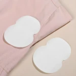 Dos parches blancos adhesivos para axilas sobre tela rosa claro y fondo beige, diseñados para absorber sudor.
