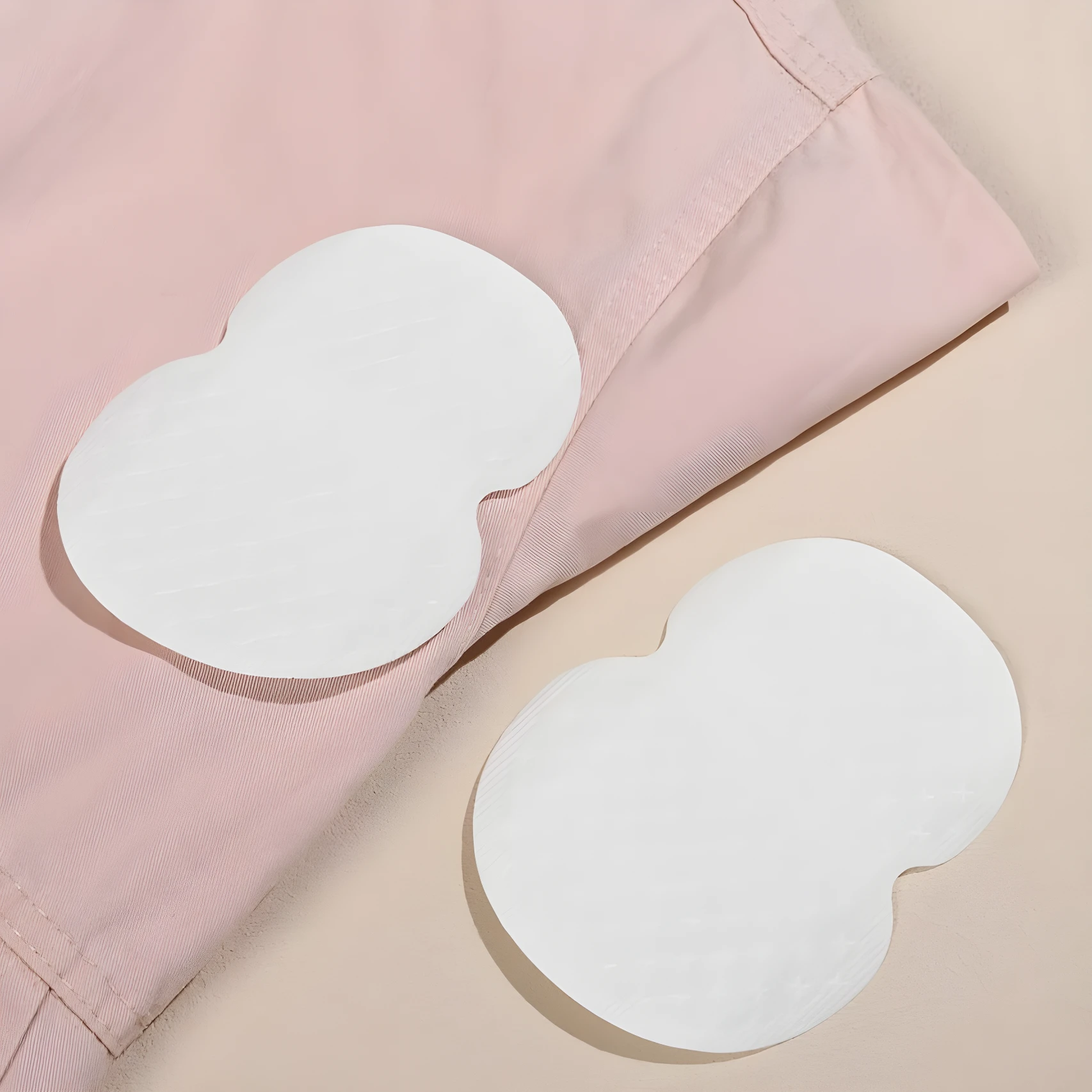 Dos parches blancos adhesivos para axilas sobre tela rosa claro y fondo beige, diseñados para absorber sudor.