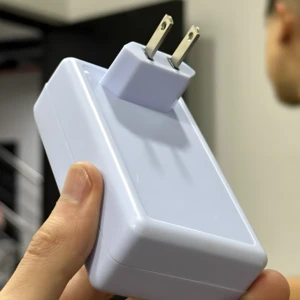 Cargador blanco, tipo pared, con clavijas planas tipo americano, sostenido en mano.