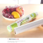 Rollos de película plástica transparente para alimentos, uno cubriendo frutas en plato y otro desenrollado, sobre mesa de madera clara.