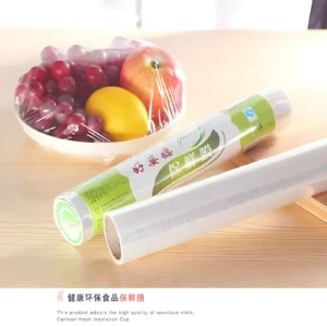 Rollos de película plástica transparente para alimentos, uno cubriendo frutas en plato y otro desenrollado, sobre mesa de madera clara.