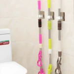 Soportes para escobas y trapeadores, uno rosa, uno verde y uno café, colgados en pared.