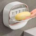 Jabonera de ducha adhesiva blanca con borde rosa, un jabón amarillo siendo colocado.