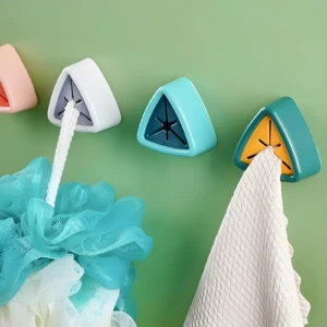 Cuatro soportes adhesivos triangulares de colores (rosa, blanco, azul claro, verde oscuro/amarillo) sujetan un paño blanco texturizado y una esponja de baño turquesa sobre pared verde.