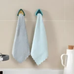 Dos paños de microfibra texturizada (uno gris y otro azul claro) colgando de soportes adhesivos triangulares verdes y azules en una pared de azulejos beige.