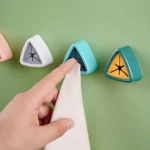 Mano insertando paño blanco texturizado en soporte adhesivo triangular azul turquesa, junto a otros soportes en blanco y verde oscuro con centro amarillo.