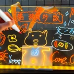 Tablero acrílico LED que muestra un cerdo sonriente, texto "Ven" y ofertas, con marcador, ideal para negocios.