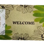Tapete felpudo atrapa polvo color beige con flores negras y hojas verdes, dice "WELCOME". Incluye etiqueta.