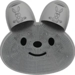 Tapete exfoliante de silicona gris con textura de cerdas, forma de cara de conejo, ranuras para pies y detalles de dibujo de conejo sonriente.