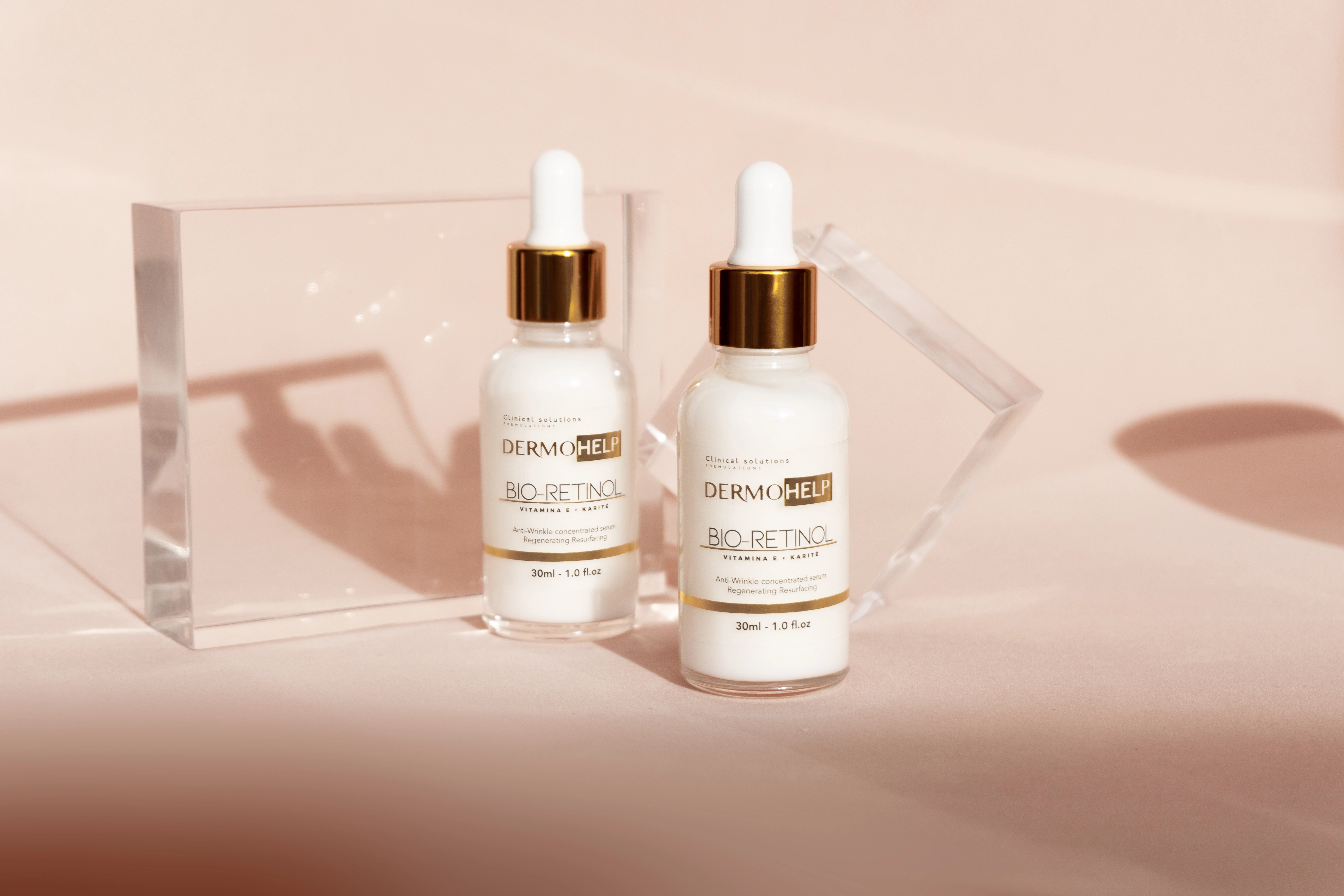 Dos frascos blancos de Sérum Bio-Retinol DermOHELP con gotero dorado, sobre fondo rosa claro, con prismas de cristal.