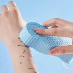 Esponja mágica exfoliante 3D azul removiendo células muertas de brazo humano con patrón escamas.