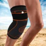 Rodillera deportiva de compresión negra con detalles naranja, usada en pierna sobre fondo de playa y cielo azul.
