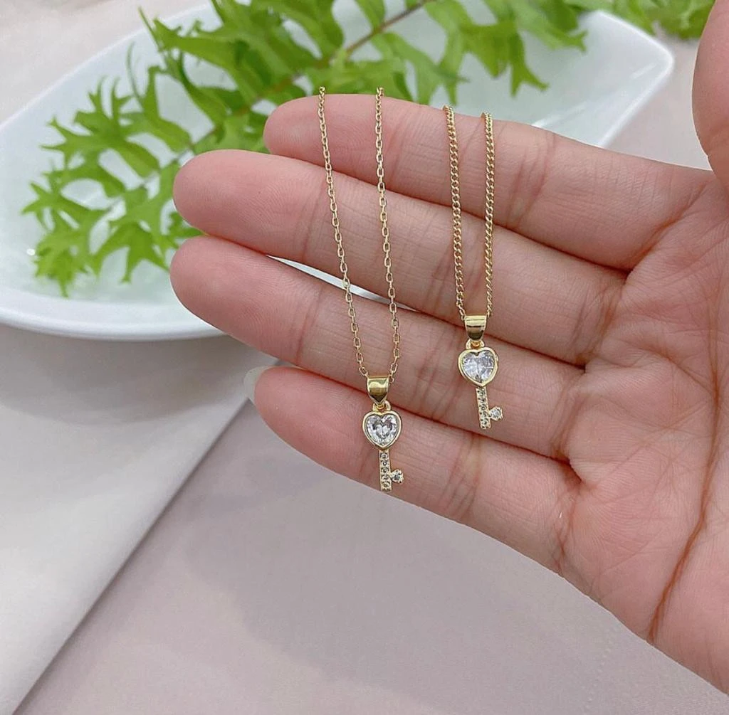 Collar de oro con dije de llave corazón y circonitas, sujetado por una mano.