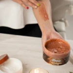 Mano aplicando exfoliante corporal de cocoa sobre el brazo. Frasco abierto a la vista.