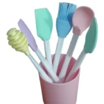 Kit de utensilios de cocina en silicona con mango blanco, incluye espátulas, cuchara, brocha y dispersor de miel en tonos pastel dentro de vaso rosa.
