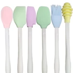 Kit de 6 utensilios de cocina en silicona con mango blanco, incluye espátulas de colores, brocha azul y dispensador de miel amarillo, dispuestos en fila.