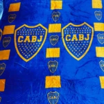 Cobija deportiva azul y dorada del CABJ, textura suave, diseño repetido con escudos y barras.