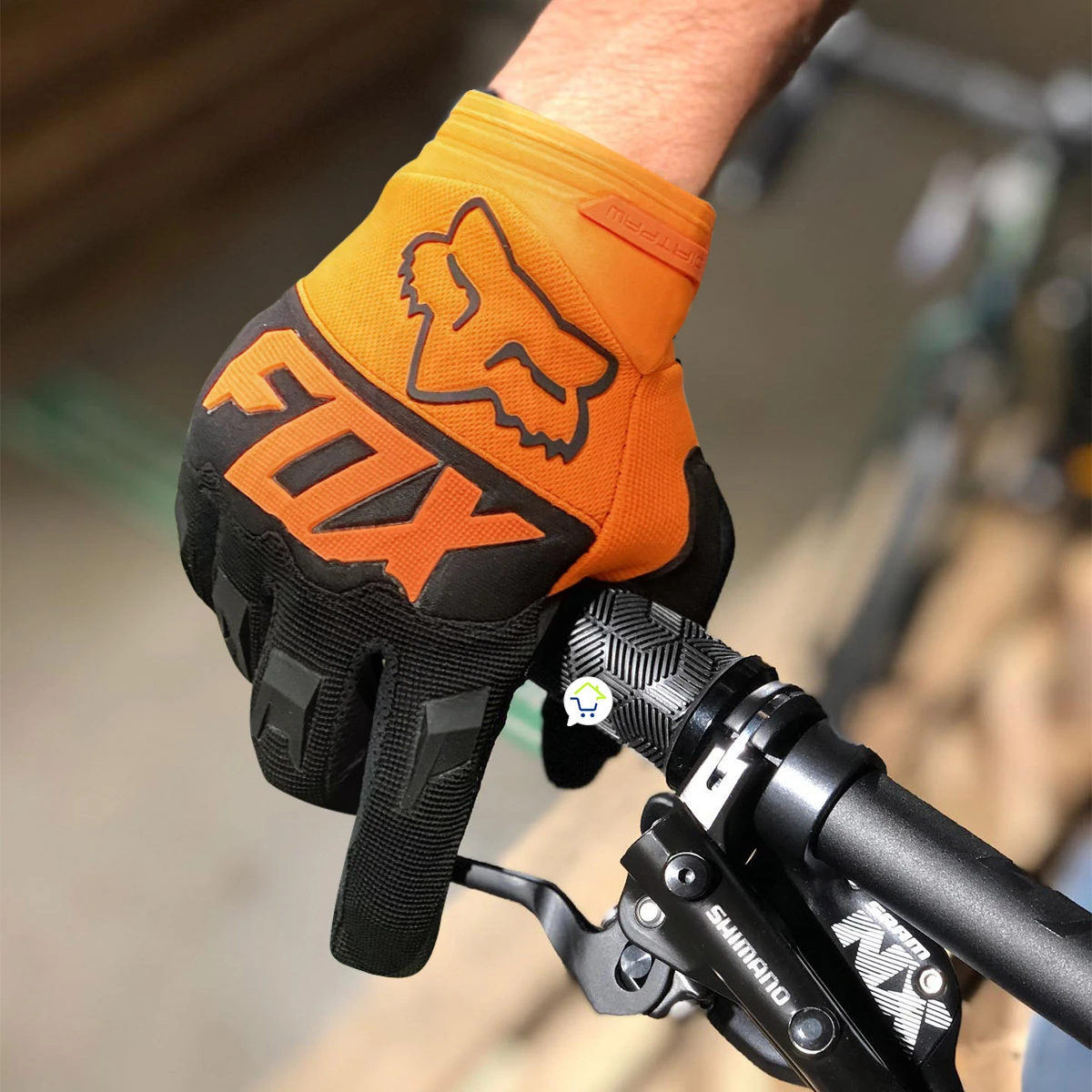 Guantes de ciclismo Fox naranja y negro con logo. Mano en manillar de bicicleta con freno Shimano.