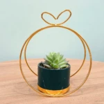 Maceta colgante dorada con forma de círculo y corazón, contiene suculenta artificial verde en maceta verde y dorada.